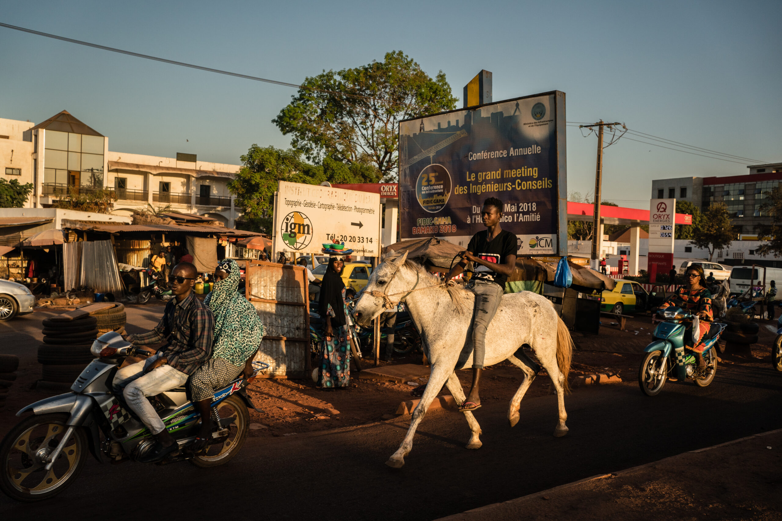 Busy street in Mali. Cars, motor-taxis, a person on a horse and pedestrians are going about their day.