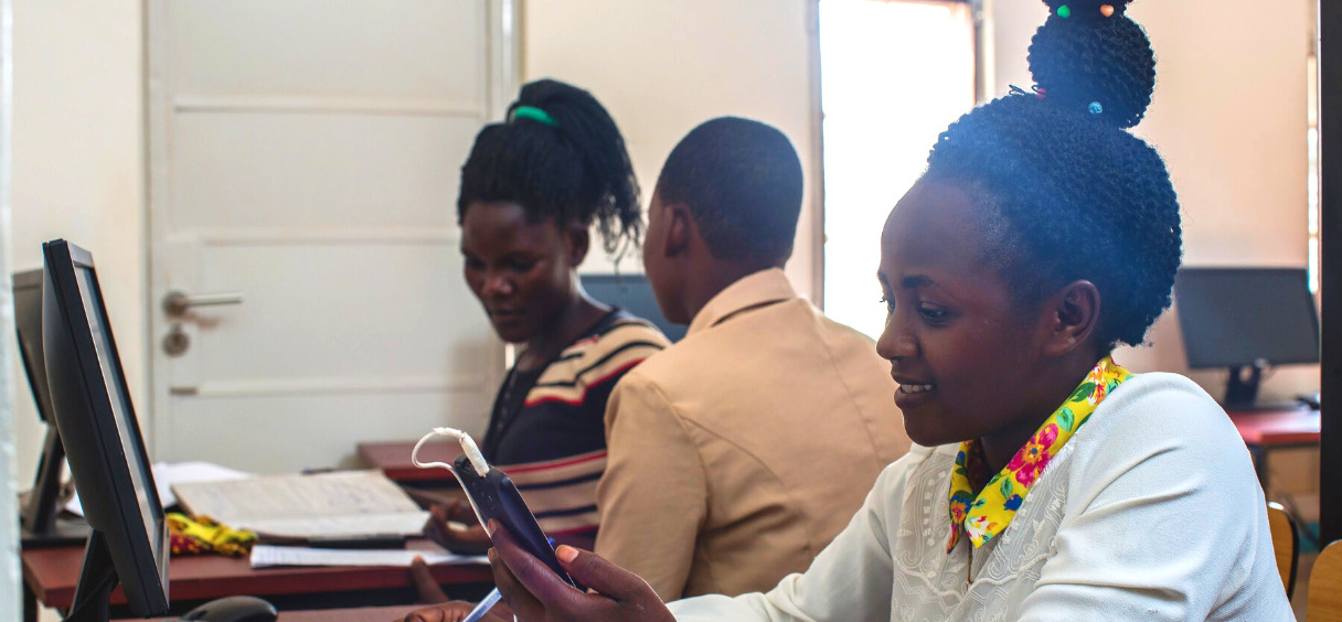 Girl sitting in a library in Uganda behind a computer, checking her smartphone.