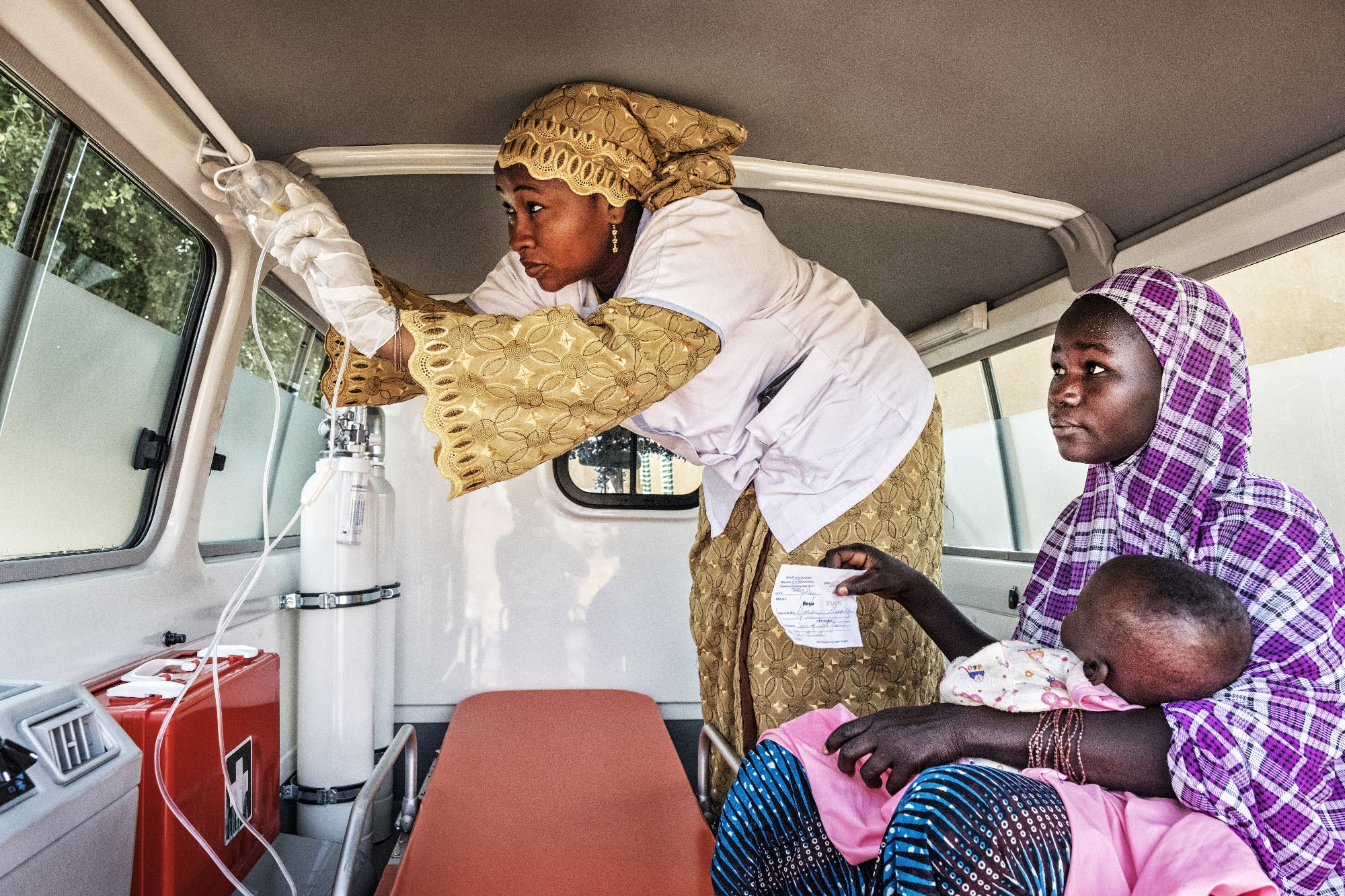 Health worker checking medical supplies in an ambulance while female patient with infant awaits.