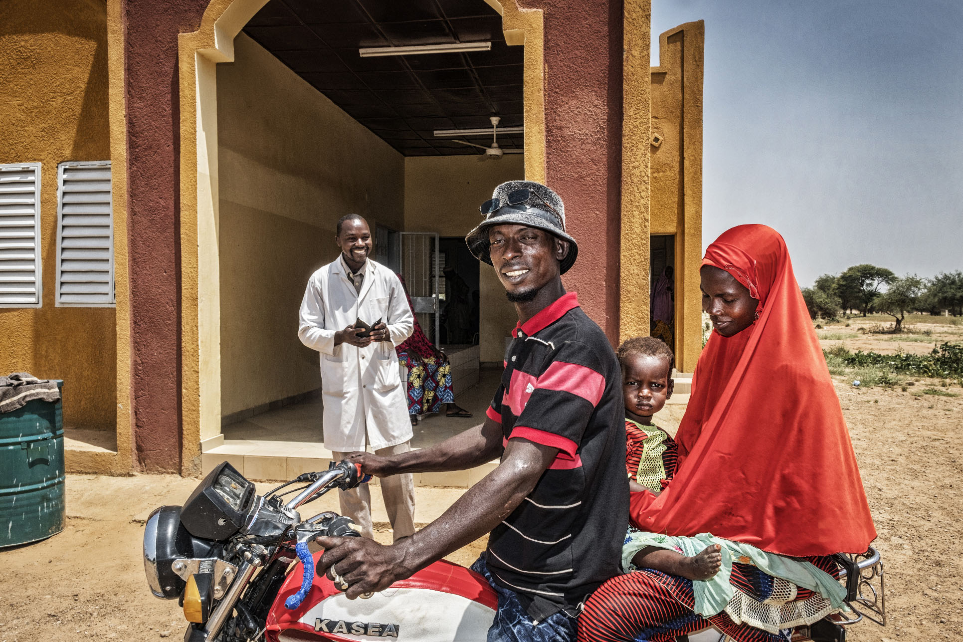 A hospital in Niger. Doctor standing outside the facility, while a woman and man arrive on a motorcycle with their baby.