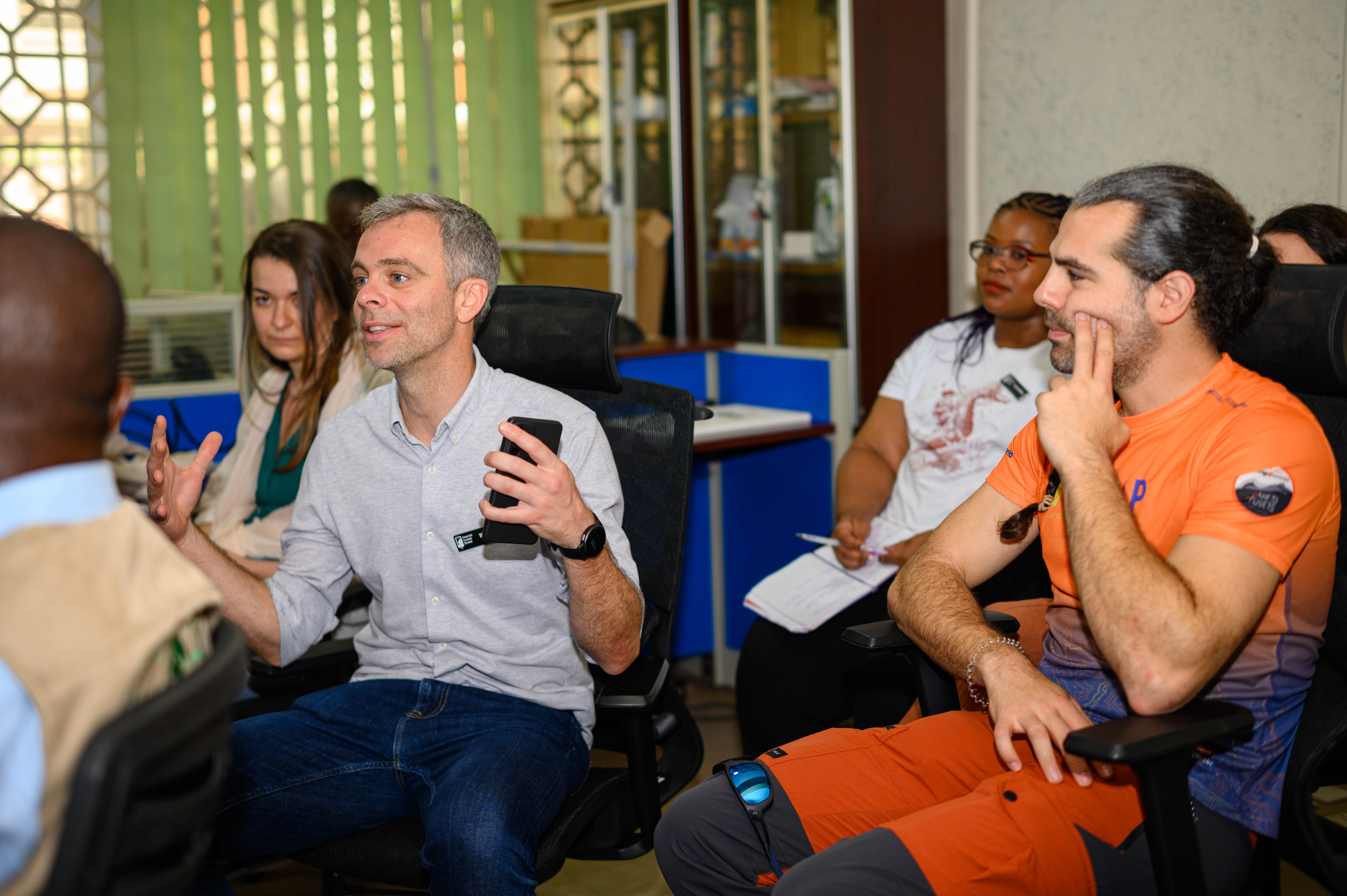One man seated, other seated people looking at him while speaking.