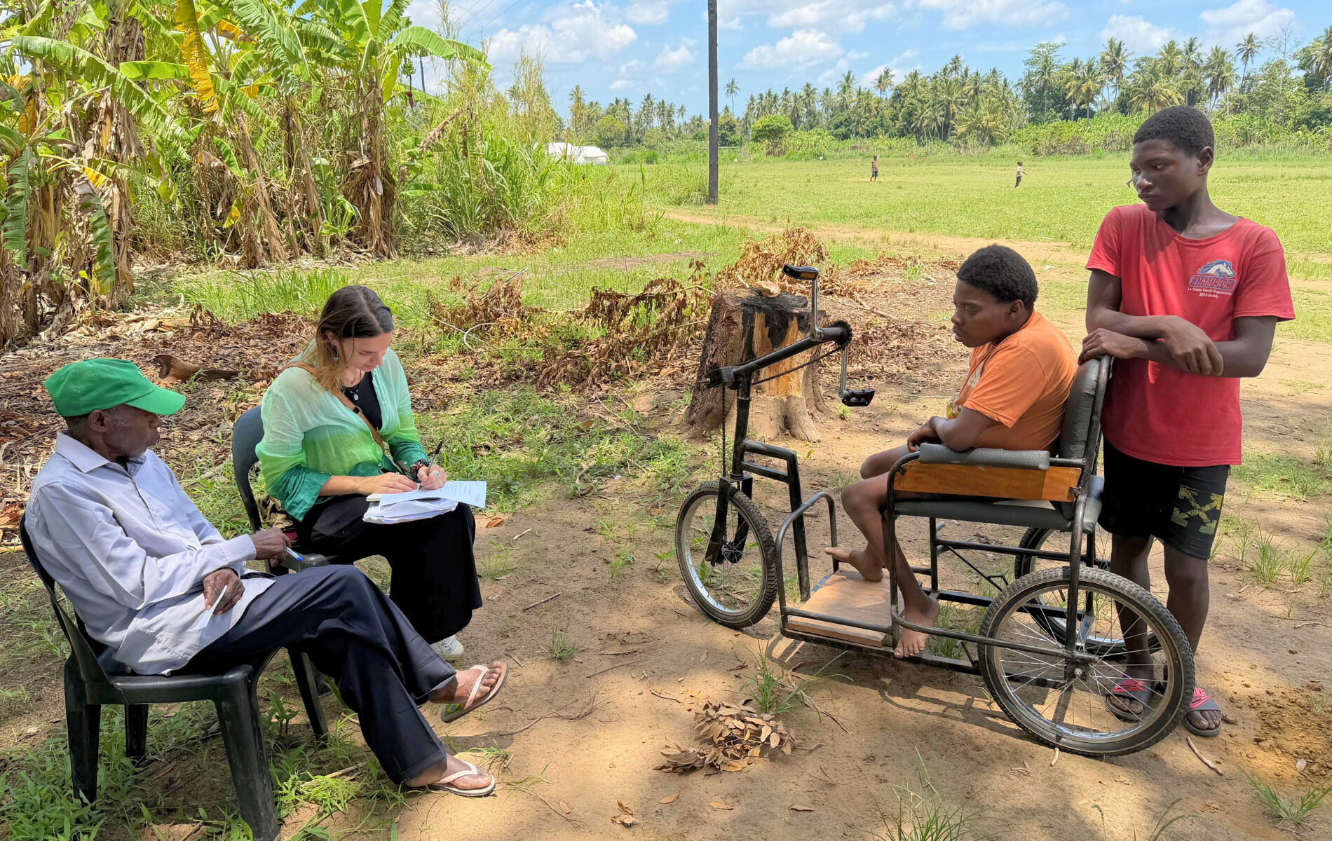 2 people sitting in a chair and writing, a person in a weelchair talking to them.