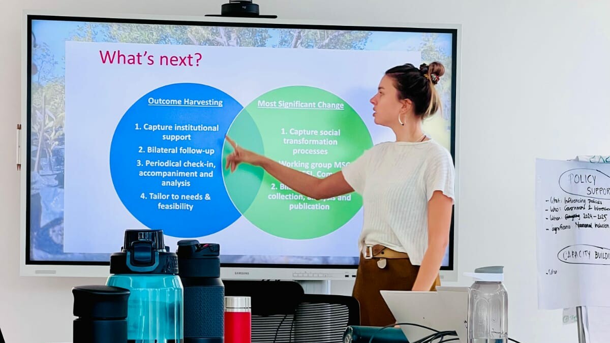 A woman stands in front of a screen, showing a powerpoint presentation.