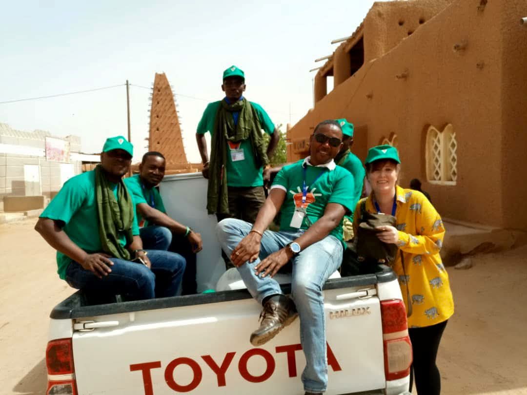 Group of young men sit in the back of a pickup truck, woman stands besides the car.