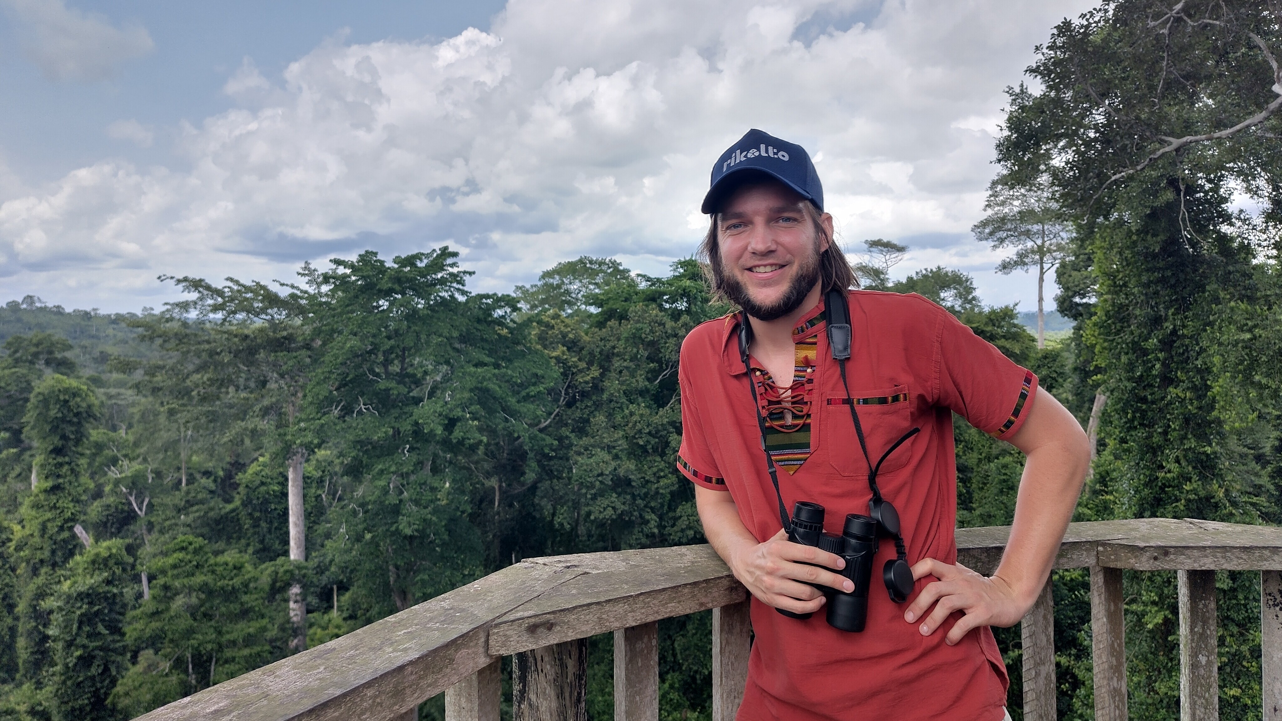 Theodore in a red T-shirt and with binoculars poses for a picture surrounded by a forest.
