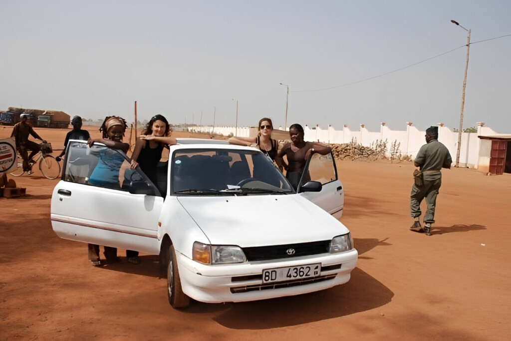 White car on the road with 4 women standing next to it.